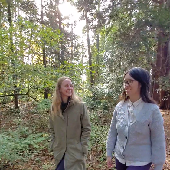 Cassandra and Juanita in the forested area by the Lower Mall Research Station. Two people walking side-by-side in a forested area. They are positioned on the middle and right side.
