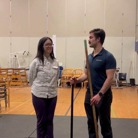 André explaining an exercise to Juanita in BodyWorks Gym. Two people are standing side-by-side in a gym. The person the left has their hands behind their back. The person on the right is leisurely holding a bar in each hand, upright.