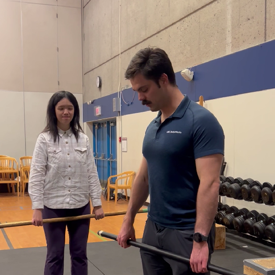 André demonstrating an exercise for Juanita in BodyWorks Gym. Two people standing in a gym. Both of them are holding a bar with both hands.